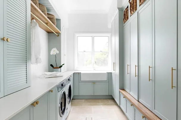 Bright and airy laundry room with light blue cabinets, washing machines, shelving, and a large window.