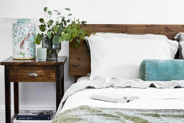 Cozy bedroom with white bedding, wooden headboard, leafy vase on nightstand, ceramic jar, and open book on bed.