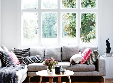 Cozy living room with a gray sofa, patterned cushions, coffee table, and large windows overlooking greenery.