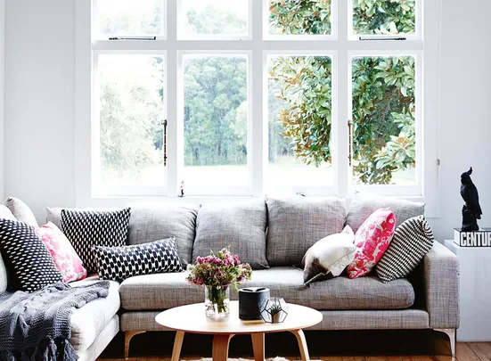 Cozy living room with a gray sofa, patterned cushions, coffee table, and large windows overlooking greenery.