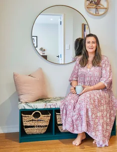 A woman in a floral dress sits on a cushioned bench holding a mug, with a round mirror and woven baskets nearby.