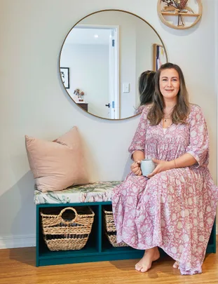 A woman in a floral dress sits on a cushioned bench holding a mug, with a round mirror and woven baskets nearby.