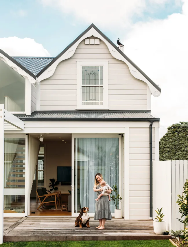 Woman holding baby stands with a dog on a wooden deck in front of a white house with a gable roof.