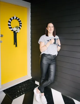 Woman in striped shirt and black pants holds mug, standing by yellow door with black-and-white wreath.