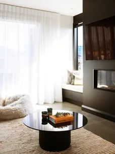 Modern living room with round table, book, and decor. Bright natural light through sheer curtains, cozy seating area.