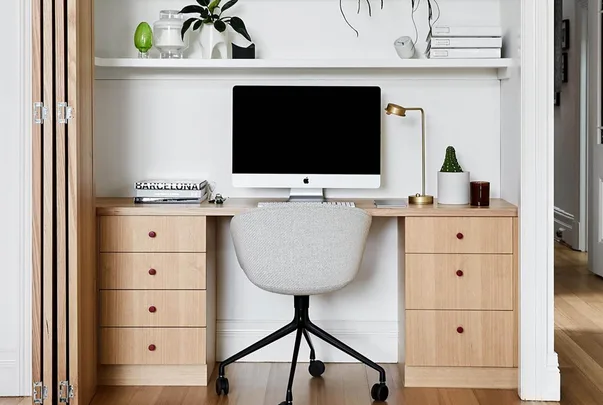 Home office with a wooden desk, computer, beige chair, gold lamp, and potted plants on shelves above.