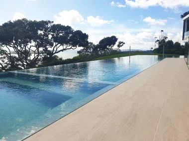 Infinity pool overlooking trees and ocean, with a clear sky and modern stone patio.