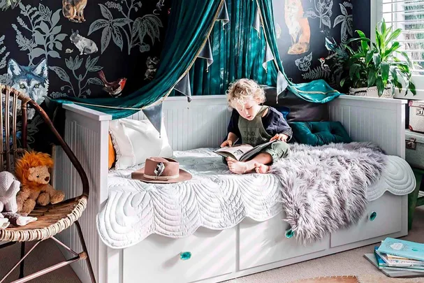 Child reading on bed with forest-themed wallpaper, plush blanket, and decorative canopy.