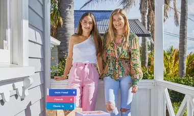 Two women smiling, standing on a porch next to stacked colorful boxes labeled "Oasis Found," "Flourish," and "Classic As."