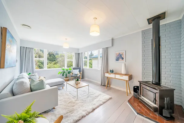 Bright living room with white sofa, fluffy rug, wood stove, and large windows showcasing greenery outside.