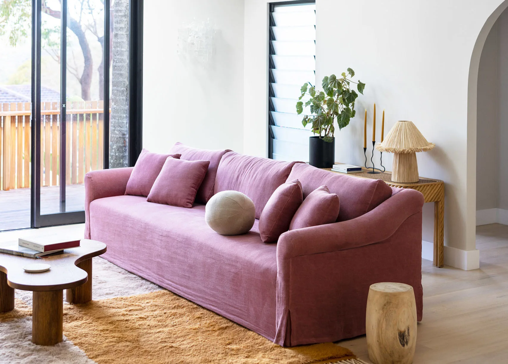 Pink sofa with cushions in a bright living room, wooden coffee table, rug, and potted plant by a window.