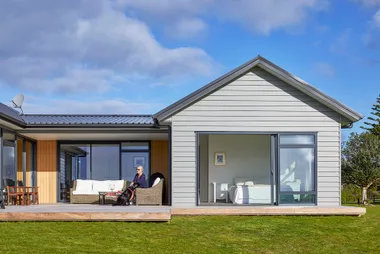 Modern house with large windows, person sitting on patio with a dog, grassy lawn under a blue sky.