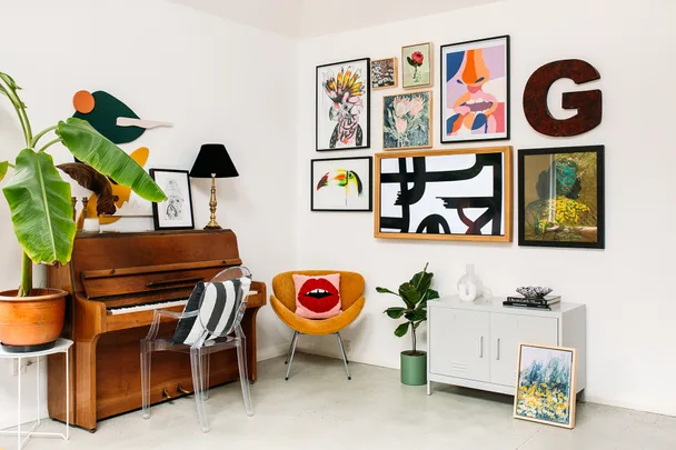 Eclectic living room with wooden piano, colorful art, clear chair, yellow chair with lip pillow, and houseplants.