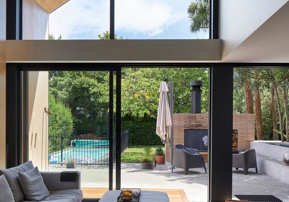 Modern living room view with large glass doors opening to a backyard patio, pool, and lush greenery.