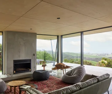 Modern living room with large windows, lush landscape view, cozy seating, rustic coffee table, and a fireplace.