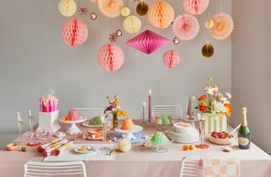 Festive dining table with colorful hanging paper decorations, flowers, champagne, desserts, and elegant tableware.