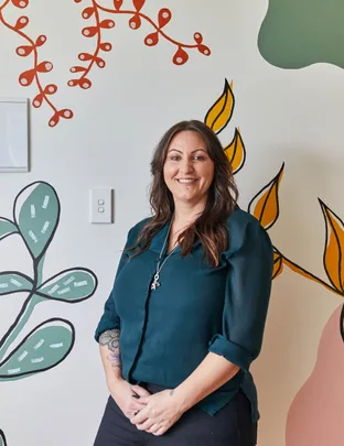 Woman smiling in front of a wall with colorful leaf and branch illustrations.
