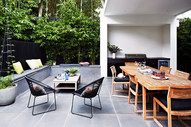 Outdoor patio with dining table, chairs, bench with cushions, plants, and a grill under a white pergola.