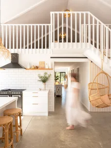 Open, airy kitchen with white cabinets, hanging chair, and a person in motion. Upper loft with stairs and slanted ceiling.