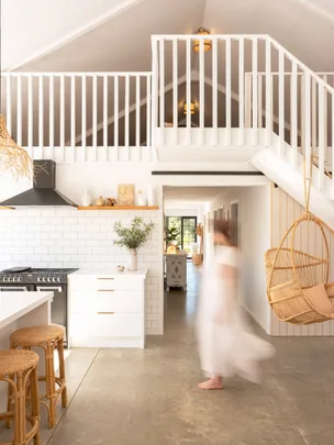 Open, airy kitchen with white cabinets, hanging chair, and a person in motion. Upper loft with stairs and slanted ceiling.