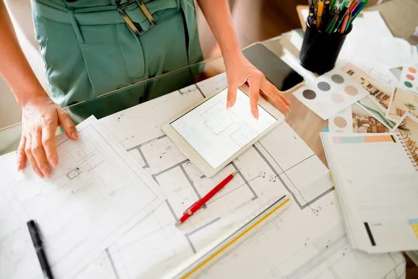 Person in green outfit reviewing architectural plans with tablet, pens, and color swatches on a desk.