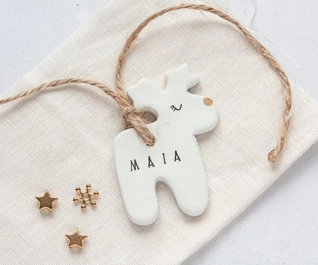 White reindeer-shaped ornament with "MAIA" text, twine, and gold star accents on fabric surface.
