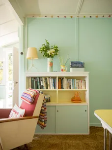 Cozy room with a pastel bookshelf, flowers, colorful blanket on a chair, books, and decorative items.