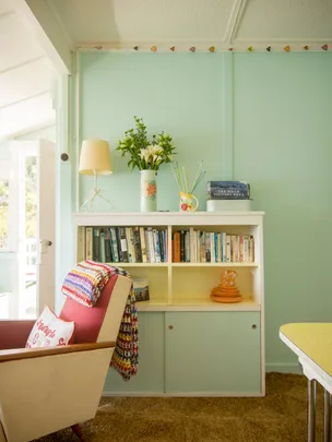 Cozy room with a pastel bookshelf, flowers, colorful blanket on a chair, books, and decorative items.