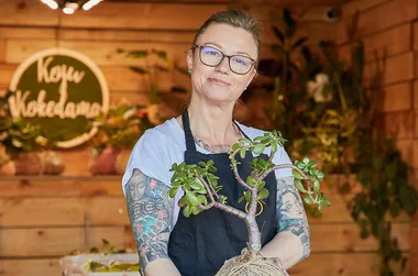 A woman with tattoos and glasses holds a potted plant in a wooden shop with "Koyu Wokedama" sign in background.