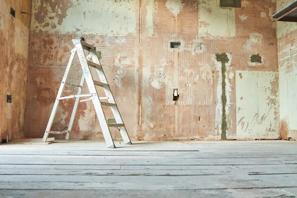 A worn, empty room with a wooden floor, unfinished walls, and a silver step ladder in the center.