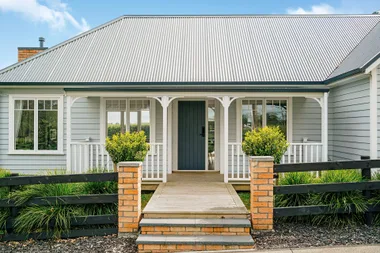 Modern bungalow with a grey corrugated roof, white railing, and a pathway flanked by brick pillars and greenery.