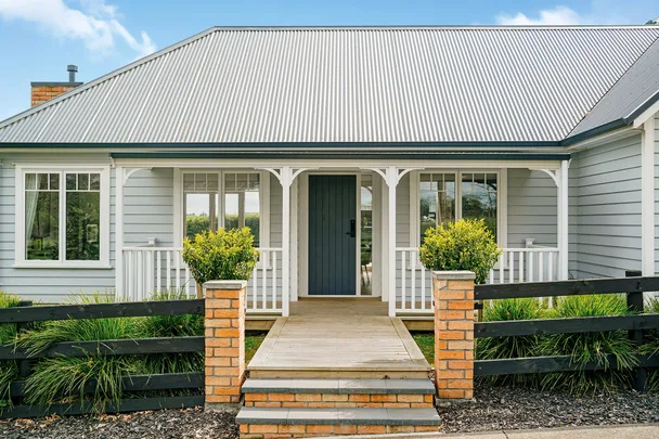 Modern bungalow with a grey corrugated roof, white railing, and a pathway flanked by brick pillars and greenery.