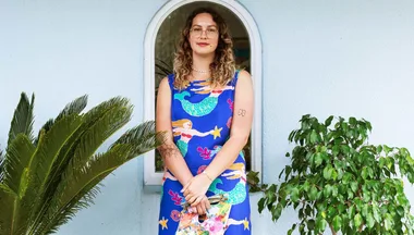 A person in a colorful mermaid-print dress stands outdoors near a window, holding a paint palette and brushes.