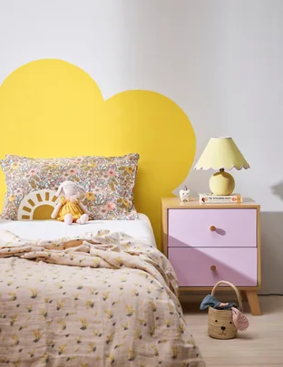 Cozy bedroom with floral bedding, stuffed bunny, yellow lamp, and pink nightstand with books and decor.