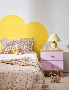 Cozy bedroom with floral bedding, stuffed bunny, yellow lamp, and pink nightstand with books and decor.