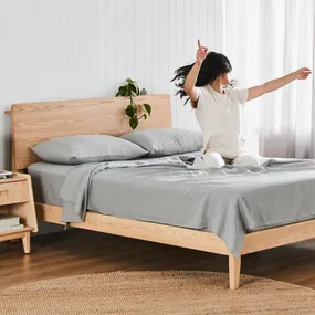 Person in white clothes joyfully stretching on a neatly made wooden bed with gray sheets in a bright room.