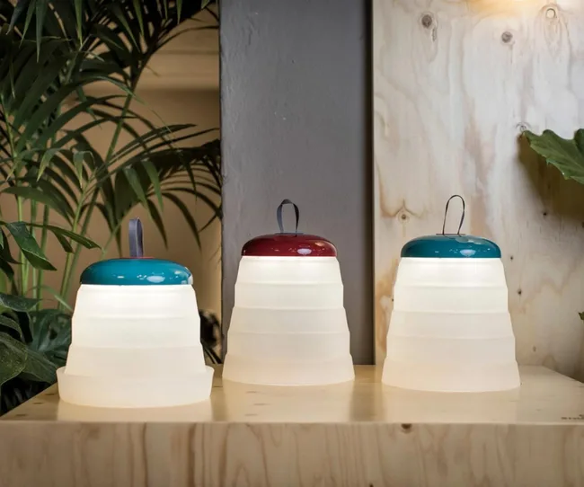 Three portable lanterns with white collapsible bodies and colorful tops on a wooden surface, surrounded by plants.