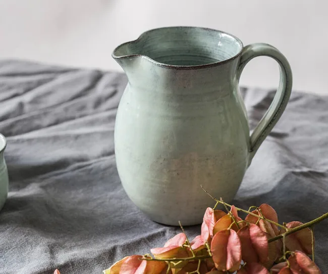 Ceramic green jug on a grey cloth with dried pink flowers nearby.