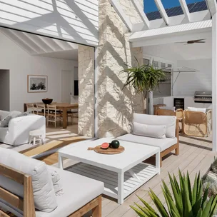 Modern indoor-outdoor living space with white furniture, exposed stone walls, and a patio featuring a coffee table and plants.