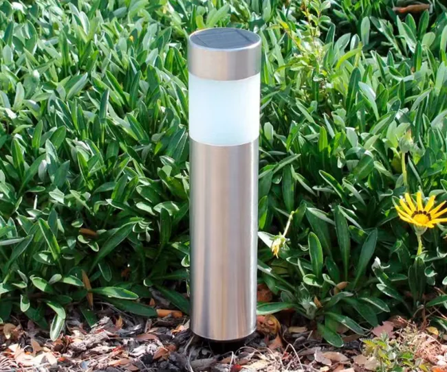 A solar garden light with a stainless steel design set amidst green foliage and a yellow flower.