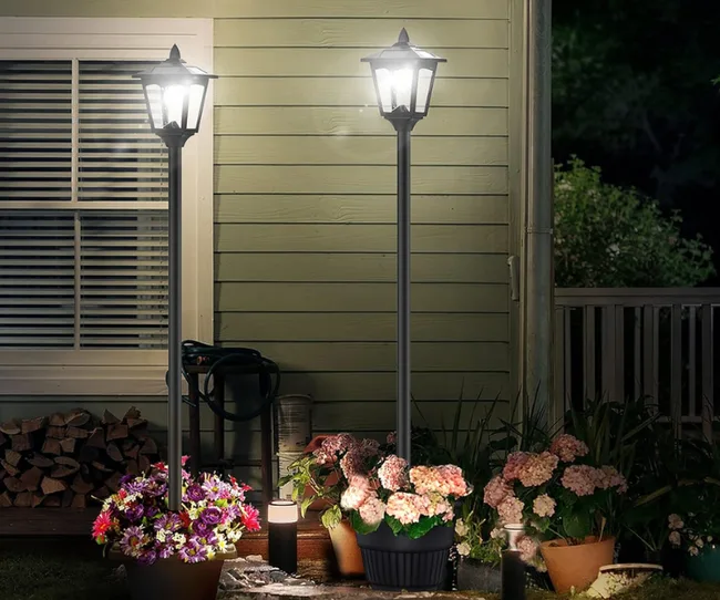 Outdoor scene with two solar lamp posts illuminating flower pots beside a house at night.