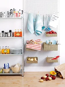 Kitchen with pegboard wall, hanging utensils, towels, metal shelves with pots, baskets of apples, and loose produce on floor.