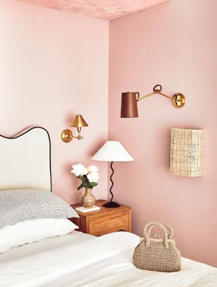 Cozy bedroom with pink walls, wooden nightstand, stylish lamps, woven wall decor, and a wicker handbag on the bed.