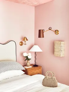 Cozy bedroom with pink walls, wooden nightstand, stylish lamps, woven wall decor, and a wicker handbag on the bed.