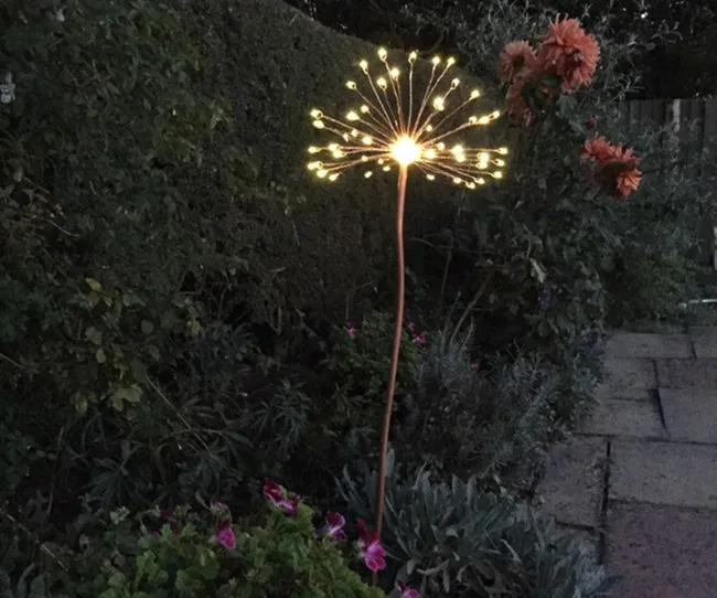 LED dandelion garden light glowing at night among plants and flowers, illuminating a garden path.