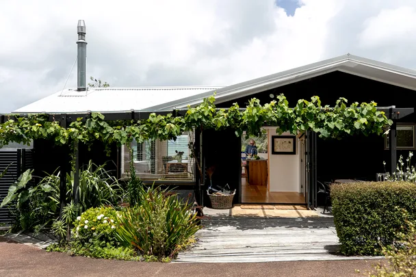 Charming house with a vine-covered pergola, lush greenery, and an open entrance revealing a cozy interior.