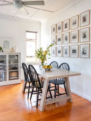 Dining room with a light wood table, black chairs, fruit bowl, wall art, and ceiling fan. White cabinet with glass doors nearby.