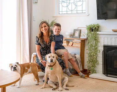 A woman and child sit in a living room with two dogs, next to a fireplace and TV, in a well-lit modern space.