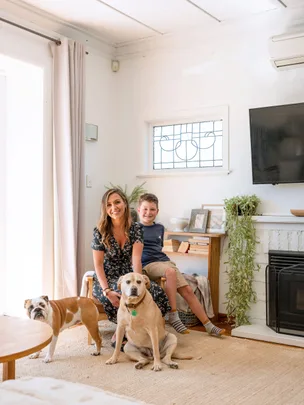 A woman and child sit in a living room with two dogs, next to a fireplace and TV, in a well-lit modern space.