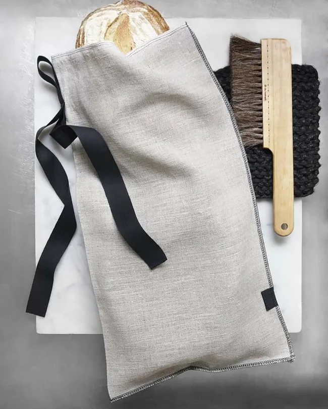 Bread in a linen bag with black ties, beside a wooden brush on a textured black mat.